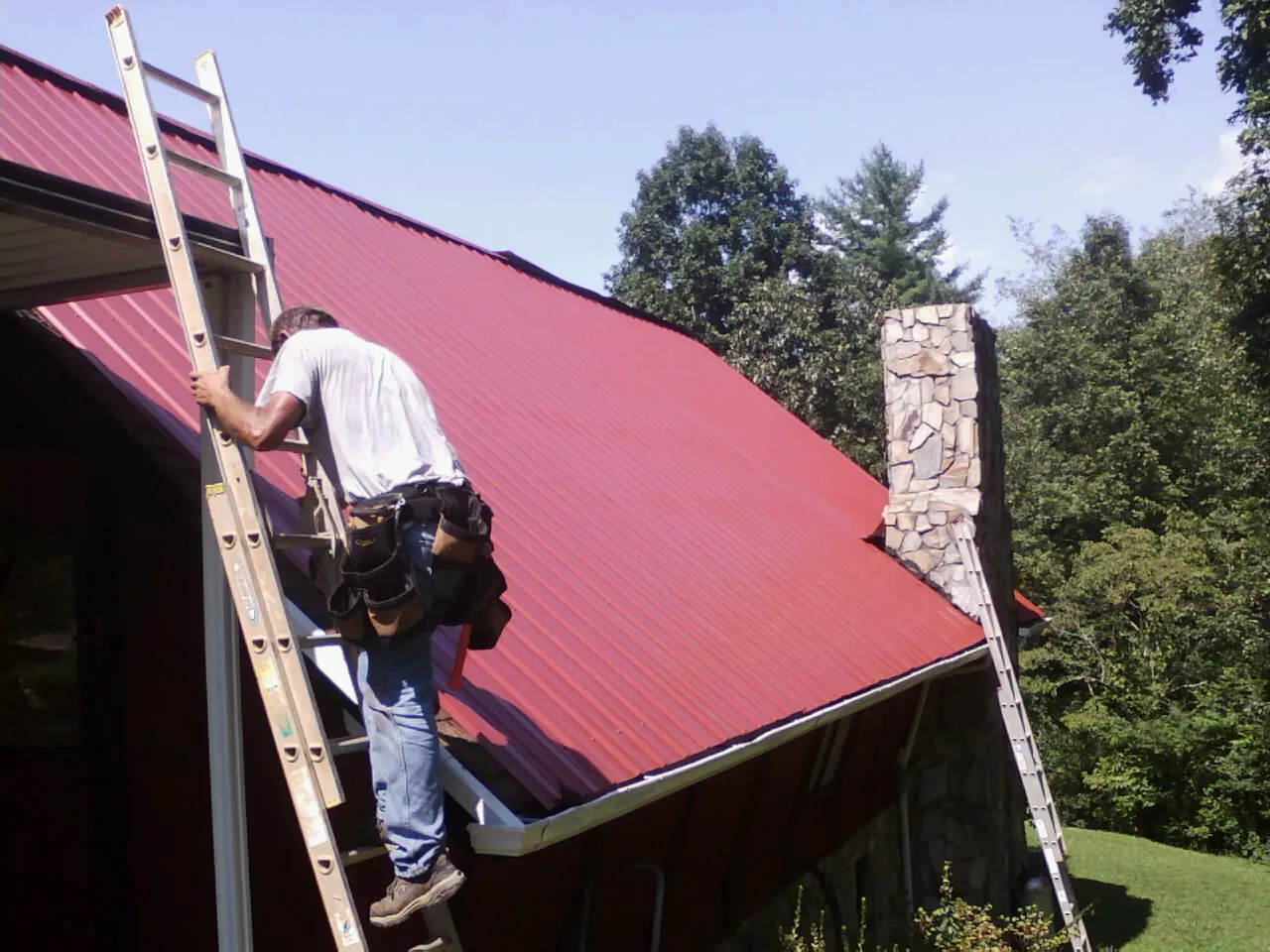 Roofer on a ladder beside a red metal roof and stone chimney — Soffit & Fascia Repair by Roofing Brothers in Wilburton